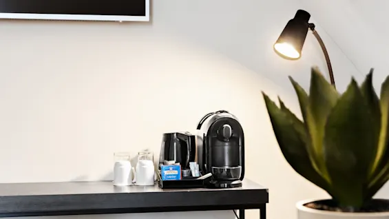 Kaffemaskine og elkedel på et sort bord med to hvide krus, glas og Twinings Lady Grey-te. I nærheden er der en lampe og en potteplante mod en lys væg.