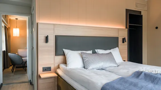 A neatly made bed with pillows and a blanket sits in a modern hotel room. Adjacent is a doorway leading to a seating area with a table and chairs.