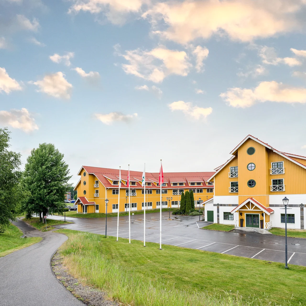 A large, yellow building with red roofs stands next to a parking lot, surrounded by trees and a pathway under a partly cloudy sky. Five flagpoles line the front.