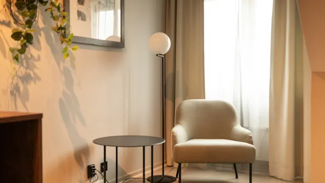 En beige lenestol står ved siden av et rundt svart bord og en høy gulvlampe i et koselig rom. Lys filtreres gjennom et vindu med lette gardiner, og en plante henger på veggen.