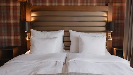 Et hyggeligt hotelværelse med en pænt redt seng, hvidt sengetøj, en polstret sengegavl og ternet tapet, klar til et behageligt ophold.