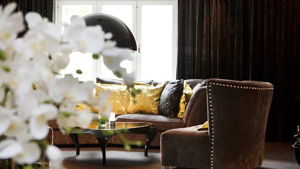 An elegantly decorated living room with plush sofas, patterned cushions, and large windows, accentuated by soft lighting and ornate flowers.
