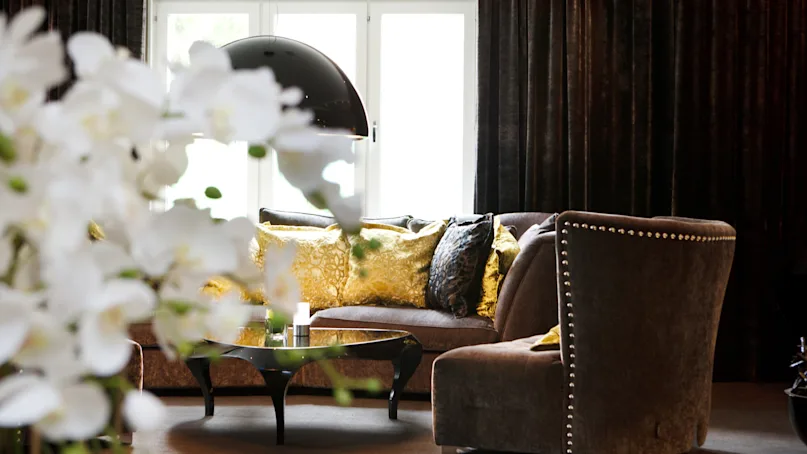 An elegantly decorated living room with plush sofas, patterned cushions, and large windows, accentuated by soft lighting and ornate flowers.
