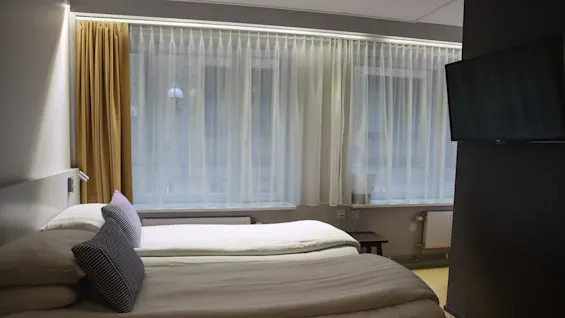 To enkeltsenger med to mønstrede puter står mot et stort vindu med gjennomsiktige gardiner. En flatskjerm-TV er montert på en mørk vegg i et koselig hotellrom.