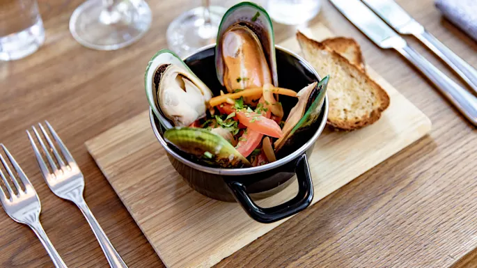 Blåskjell i en svart gryte står på en treplate, pyntet med urter og skivede grønnsaker, sammen med to brødskiver. Bestikk og vinglass ses på trebordet.