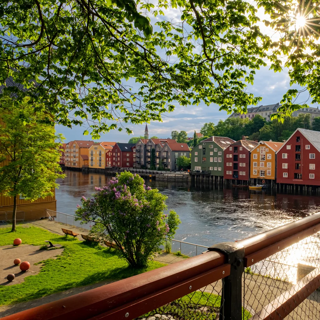 Fargerike hus langs en elv under en lys himmel, innrammet av grønne blader. En park med benker ligger ved vannet.