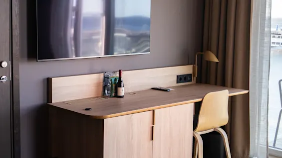 Et skrivebord i tre har en vinflaske, snacks og en fjernkontroll. En flatskjerm-TV henger over, med gardiner og et vindu i nærheten, og skaper en hotellrom-lignende setting.