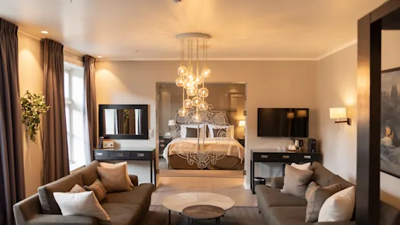 A cozy room features a central bed adorned with elegant bedding, surrounded by two sofas and a round coffee table. Warm lighting and decorations add to the inviting, stylish atmosphere.