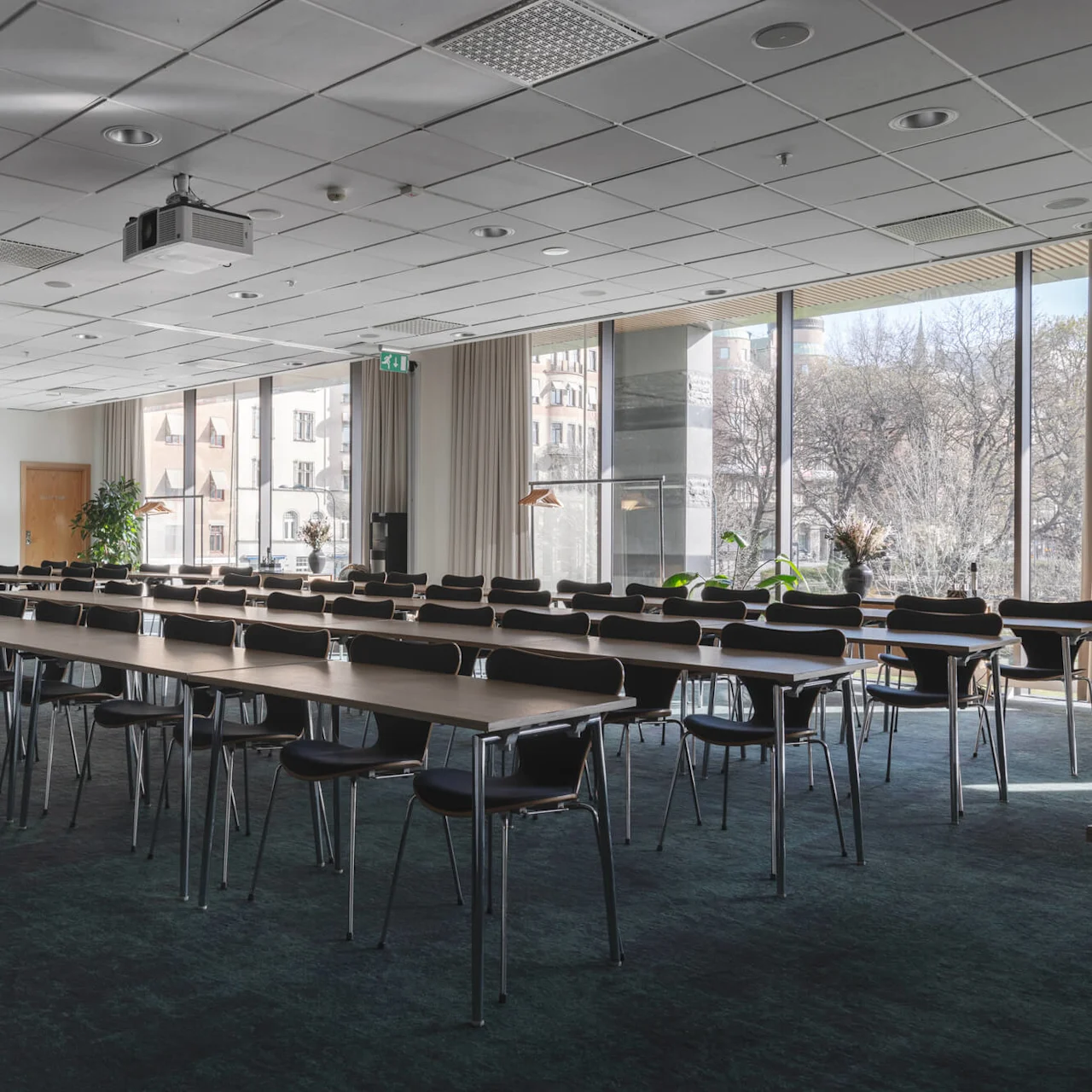 Bord och stolar är ordnade i rader i ett ljust konferensrum med stora fönster som visar en stadsutsikt utanför. Projektorer och växter dekorerar rummet. Utgångsskylt synlig.