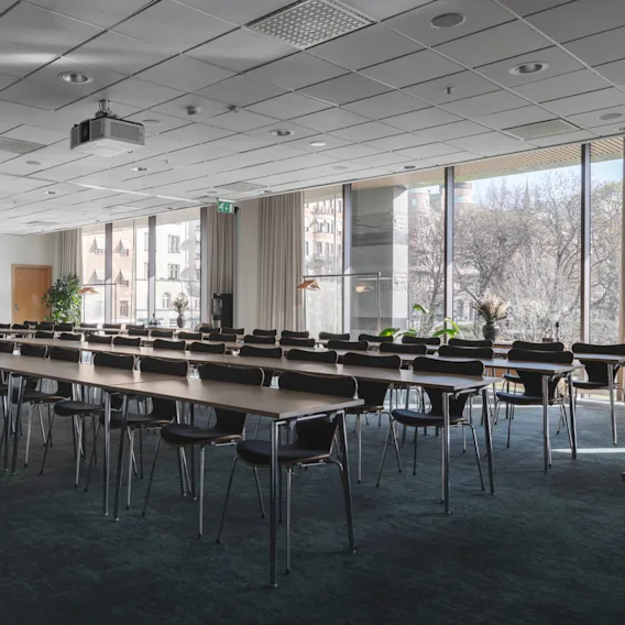Bord och stolar är ordnade i rader i ett ljust konferensrum med stora fönster som visar en stadsutsikt utanför. Projektorer och växter dekorerar rummet. Utgångsskylt synlig.