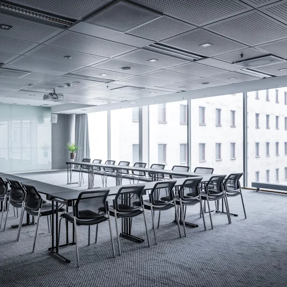 Conference room setup with a U-shaped table arrangement surrounded by chairs. Tall windows on the side provide natural light, and potted plants decorate the corners.