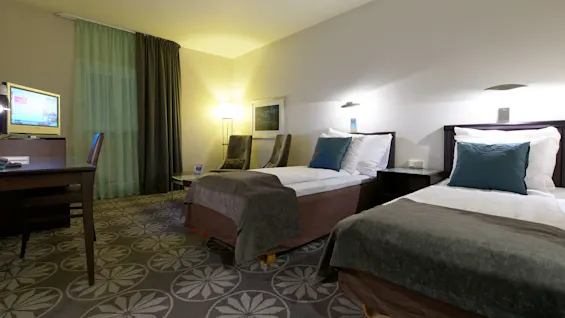 A twin hotel room with neatly made beds, nightstands, a desk with a TV and chair. The room is well-lit, with a patterned carpet and warm tones.