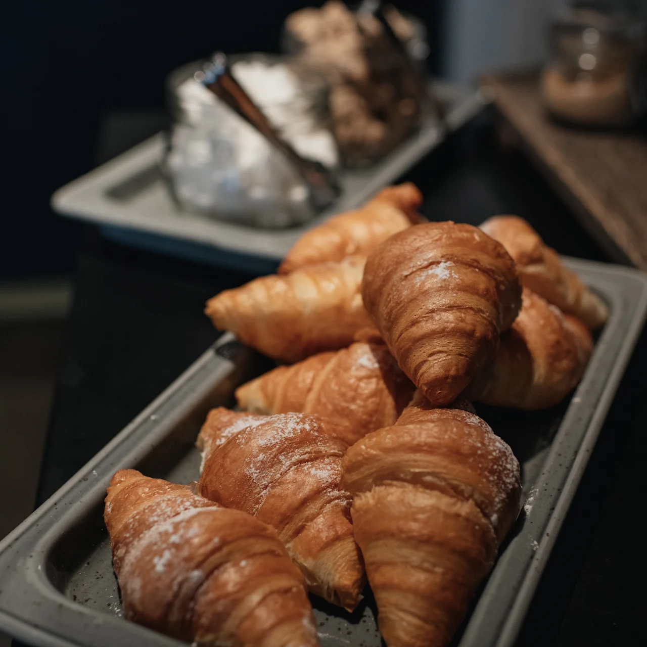 Croissants are arranged on a tray, dusted with powdered sugar. They rest on a dark table in a cozy setting, with additional baked goods in the background.