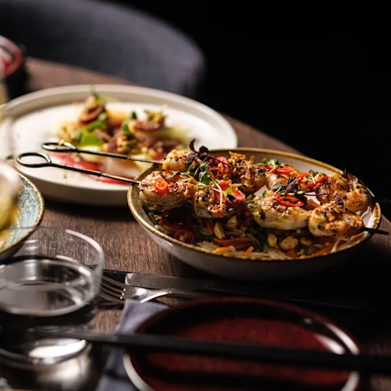 Grilled shrimp skewers rest on a bed of vegetables and rice, garnished with herbs and peppers, plated on a wooden table with wine glasses and cutlery nearby, in a dimly lit setting.