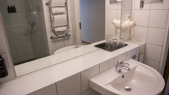 A white sink with a silver faucet sits on a tiled counter, adjacent to a large mirror reflecting a towel rack and shower; a hairdryer and glasses are nearby.
