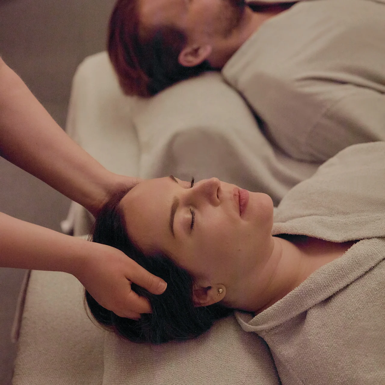 Two people lie on massage tables wearing robes, a hand massages one person's head. The setting is a serene spa environment.