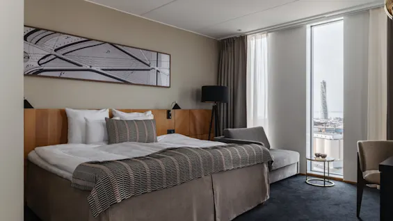 Et moderne hotellrom med en stor seng, en sjeselong og et gulv-til-tak-vindu som tilbyr utsikt over byen.