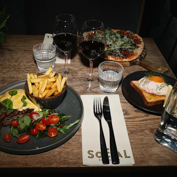 A restaurant meal on a wooden table featuring steak, fries, pizza, toast, and glasses of red wine.