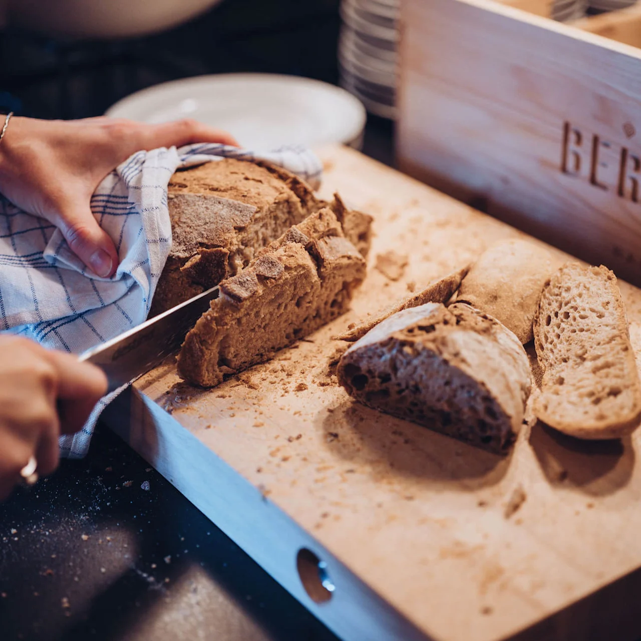 A person cuts crusty bread on a wooden board with a cloth holding the loaf. "BRØDRE BERGUS" is carved into a box in the background.