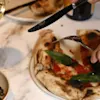 En person skærer en pizza med kniv og gaffel på et marmorbord. Nærved er der et glas hvidvin og en ret med ost og grønne blade.