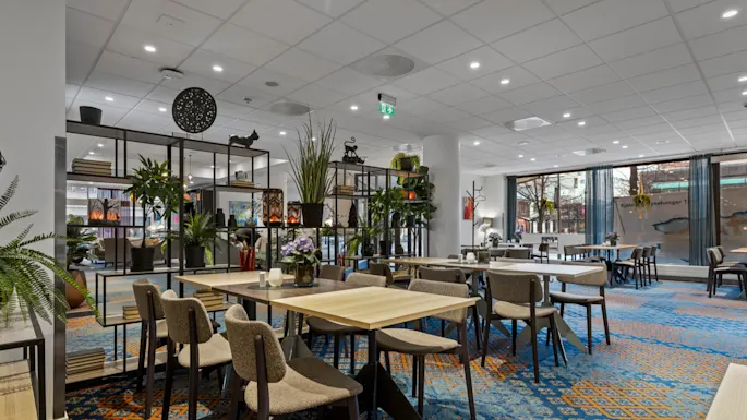 Et romslig hotellspiseområde med flere bord og stoler, dekorert med planter og hyller. Store vinduer vender ut mot en gate. Kjole rsebenger 1.