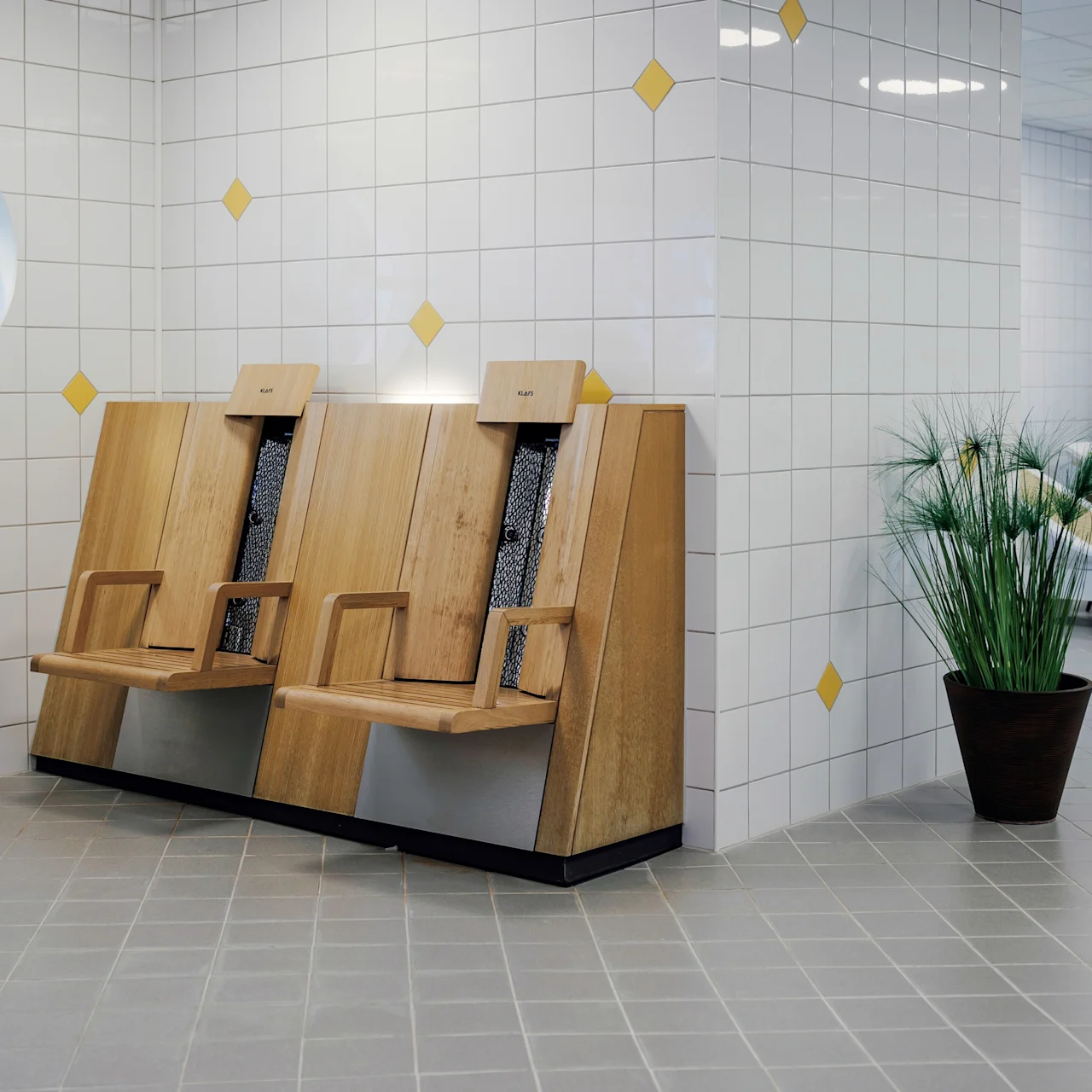 Wooden massage chairs stand by a tiled wall adorned with yellow diamonds, surrounded by plants and near lounge chairs, creating a relaxing spa-like atmosphere.