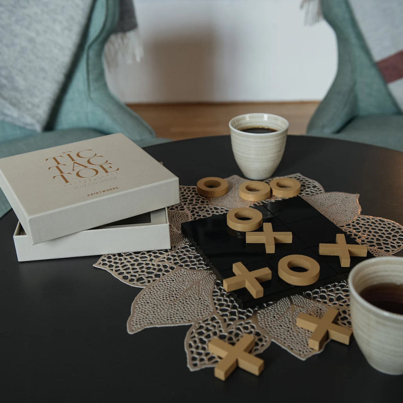 A tic-tac-toe game with wooden X and O pieces is laid out on a black board, set on a round table between two green chairs. Two cups of coffee are nearby.