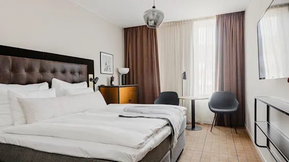 En seng med hvitt sengetøy og en mørk sengegavl i et moderne soverom. En lampe og kunstverk pryder et sidebord, med stoler og et bord i nærheten, delvis tildekket vindu.