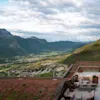 Terrasse med bord og stoler med utsikt over en grønn dal omgitt av høye fjell under overskyet himmel.