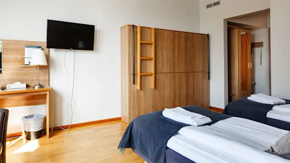 Ett hotellrum med två enkelsängar prydligt arrangerade med vikta handdukar, ett träskåp med en stege, en väggmonterad TV och ett skrivbord med en lampa och papperskorg i närheten.
