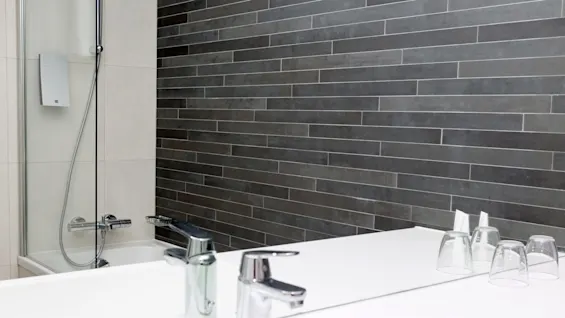 Vask med moderne metallkran, stasjonær, på hvit benk ved siden av omvendte glass ved siden av et glassinnfattet badekar i et bad med grå fliser.