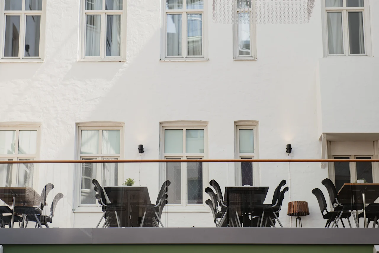 Utomhus sittplats med flera bord och stolar, placerade på en terrass. Omgiven av vita byggnadsväggar med fönster. Lugn atmosfär.