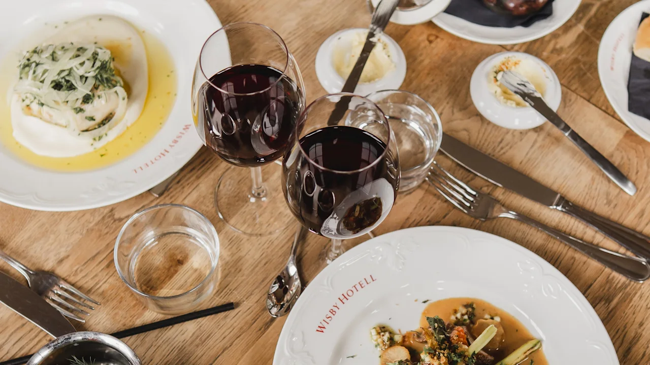 Norwegian Bokmål: To tallerkener med gourmetmat står på et trebord, akkompagnert av rødvinsglass og små boller, i en elegant restaurantinnstilling.