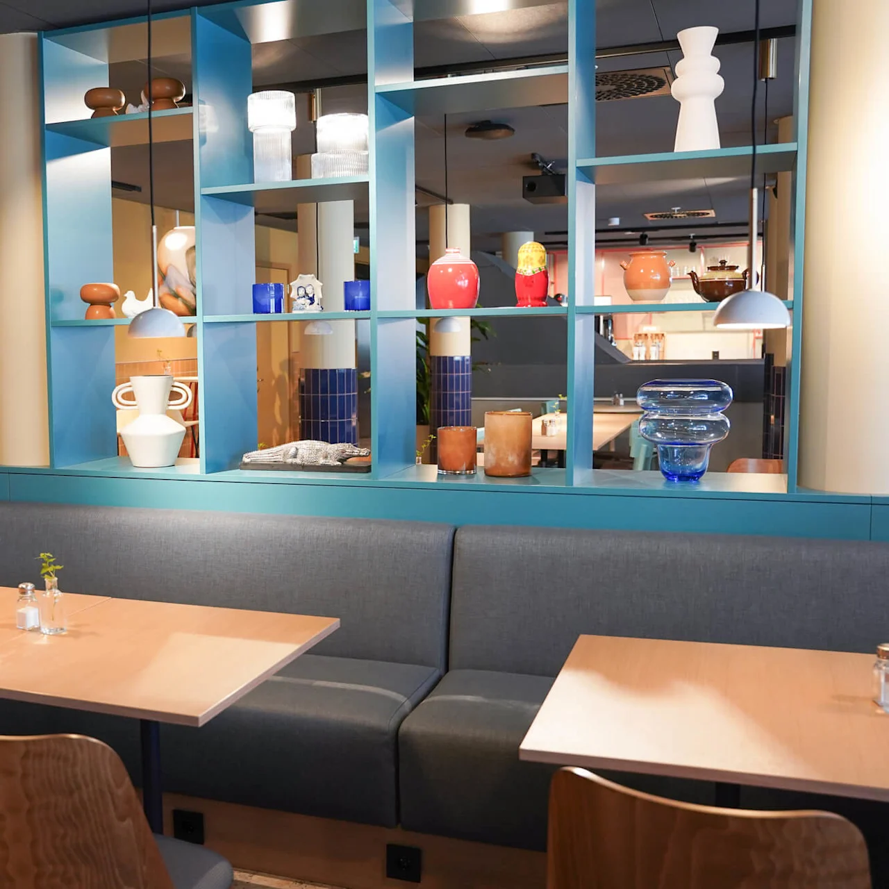 Dining tables with wood chairs and small plants; a blue shelving unit displays colorful pottery and glassware; softly-lit modern cafe with beige walls and hanging lamps.