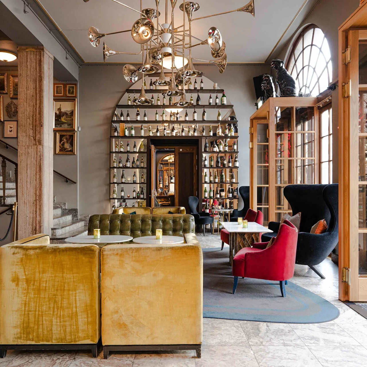A brass chandelier hangs above a stylish lounge with velvet seating and tables. Shelves filled with bottles line a wall, surrounded by decorative paintings and wooden doors.