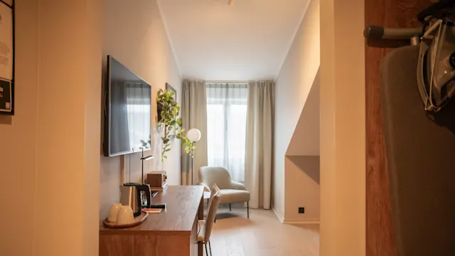Et trebord holder en lampe og vannkoker; en stol og TV er i nærheten. Rommet har lyse vegger, tregulv og vinduer med tynne gardiner, og skaper en varm, godt opplyst atmosfære.