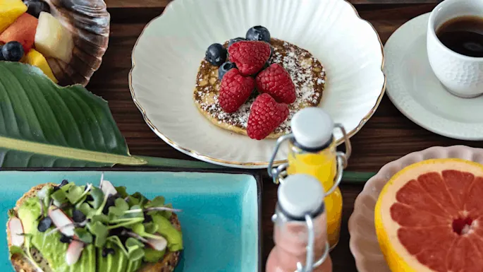 A round pancake topped with raspberries and blueberries sits on a white plate beside a cup of coffee. Nearby, avocado toast and sliced grapefruit complement the breakfast table.