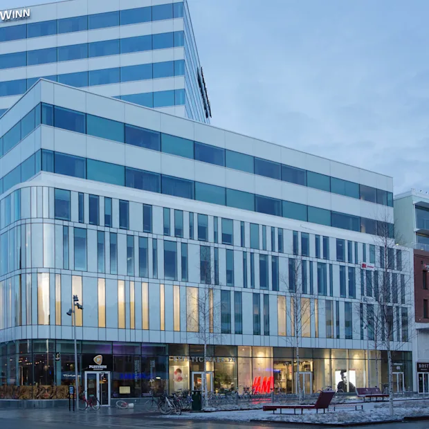Nykyajan monikerroksinen hotellirakennus, lasijulkisivu, valaistu sisätila, iltahämärän taivas.