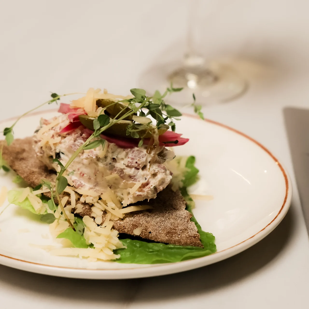 A dish of bread topped with creamy salad, pickled vegetables, microgreens, and grated cheese on a lettuce leaf. Positioned on a white plate beside a wine glass and cutlery.