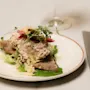 A dish of bread topped with creamy salad, pickled vegetables, microgreens, and grated cheese on a lettuce leaf. Positioned on a white plate beside a wine glass and cutlery.