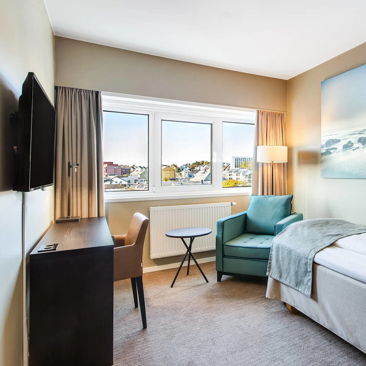Et ryddig hotellrom viser en enkeltseng med turkise detaljer. Et maleri av en kystscene pryder veggen. Et vindu viser en urban byutsikt under en klar himmel.