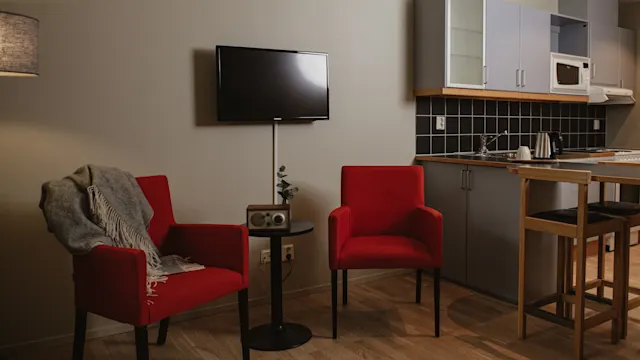 Två röda fåtöljer är placerade i ett vardagsrum med ett modernt kök i bakgrunden. Ett litet bord mellan fåtöljerna håller en radio under en väggmonterad TV. Mjukt ljus tillför värme.