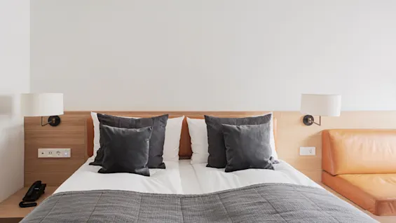 Double bed with gray pillows and blanket, neatly arranged against a wooden headboard. Two wall lamps are mounted beside it. A tan leather couch is nearby, in a minimalistic room.