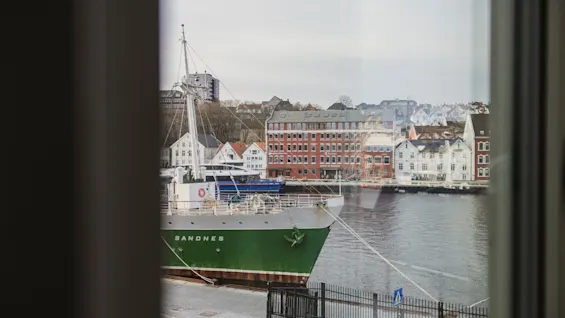 En naturskøn havneudsigt fra et vindue, med et grønt og hvidt skib ved navn SANDNES fortøjet ved en charmerende by.