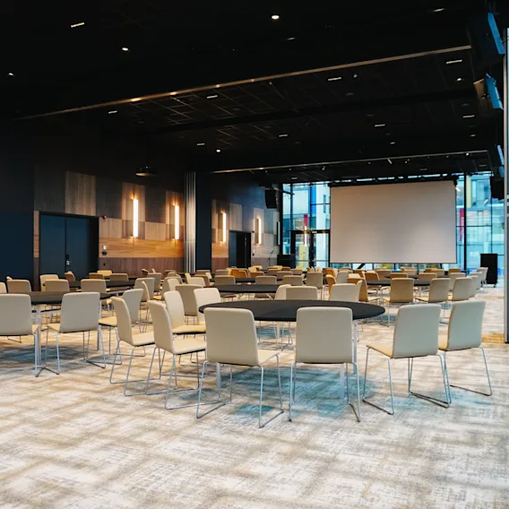 Rader av beige stoler omgir runde bord i et romslig, svakt opplyst konferanserom med trevegger og en stor projeksjonsskjerm foran.