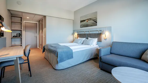 A well-lit hotel room features a neatly made double bed, a gray sofa, a desk with a lamp, and a wall painting, creating a cozy atmosphere.