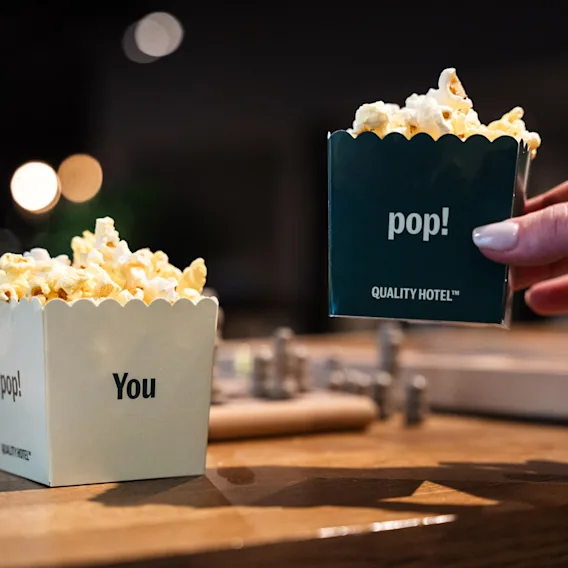 En hand sträcker sig efter en popcornlåda märkt 'pop!' medan en annan låda märkt 'pop! You' står på ett träbord.