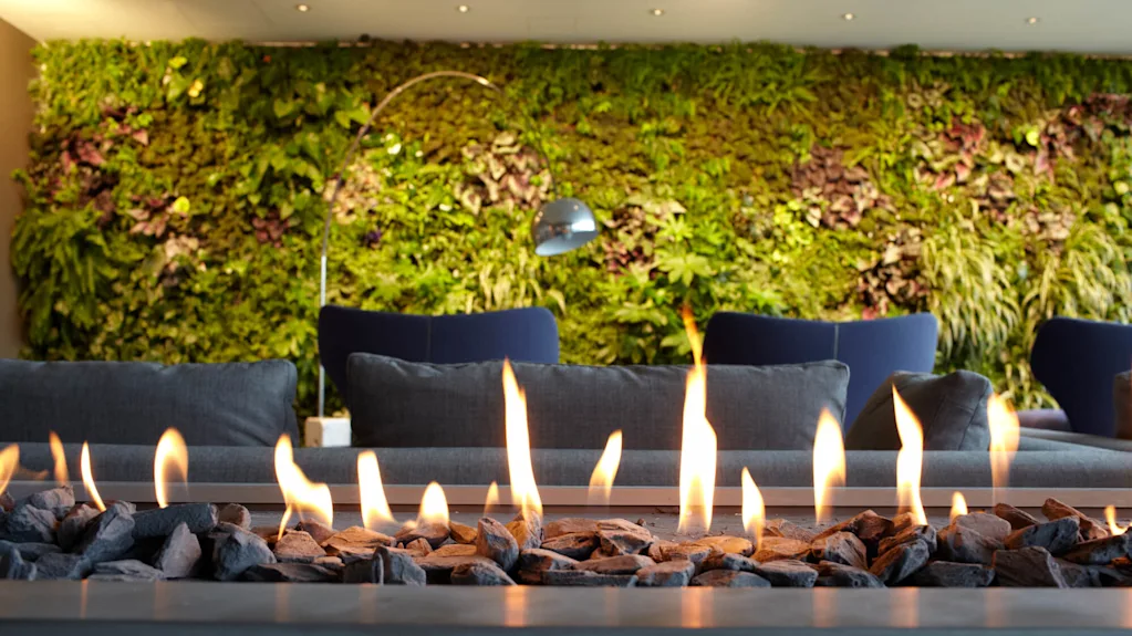 Peis med flammer, omgitt av steiner, plassert foran blå sofaer. En frodig, grønn vertikal hagevegg er i bakgrunnen, som skaper en koselig, moderne atmosfære.