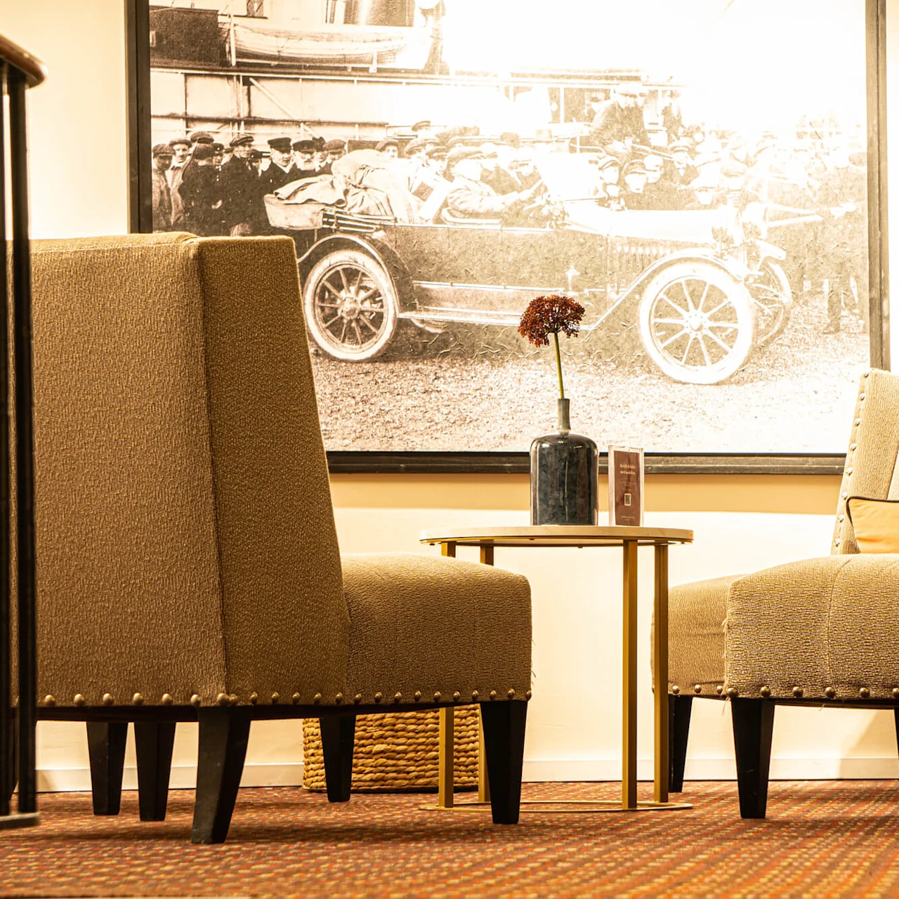 Two upholstered armchairs flank a small table with a vase, against a backdrop of a vintage car photograph and a potted plant.