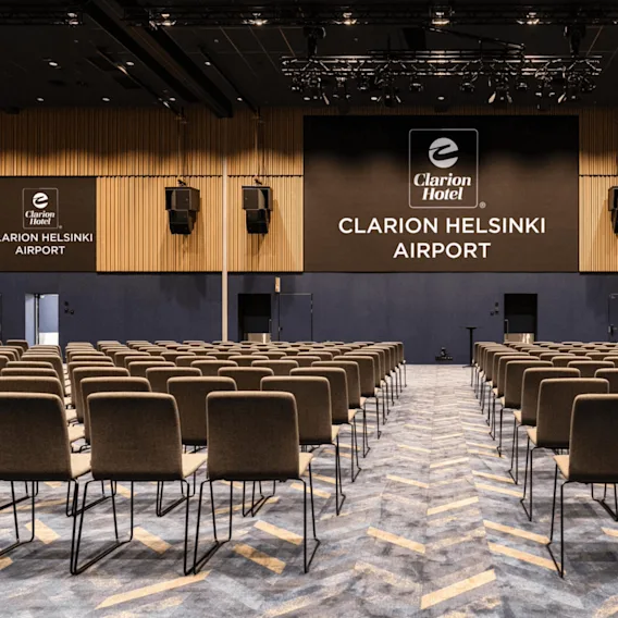 Rivien tyhjiä tuoleja on suunnattu kohti suurta näyttöä, jossa lukee "Clarion Hotel, Clarion Helsinki Airport," modernissa kokoussalissa, jossa on kuvioitu matto ja kattovalot.